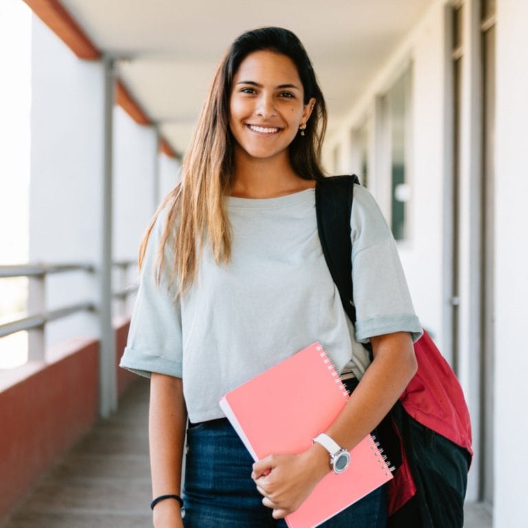 High school student standing in a school hallway holding a notebook and wearing her bookbag